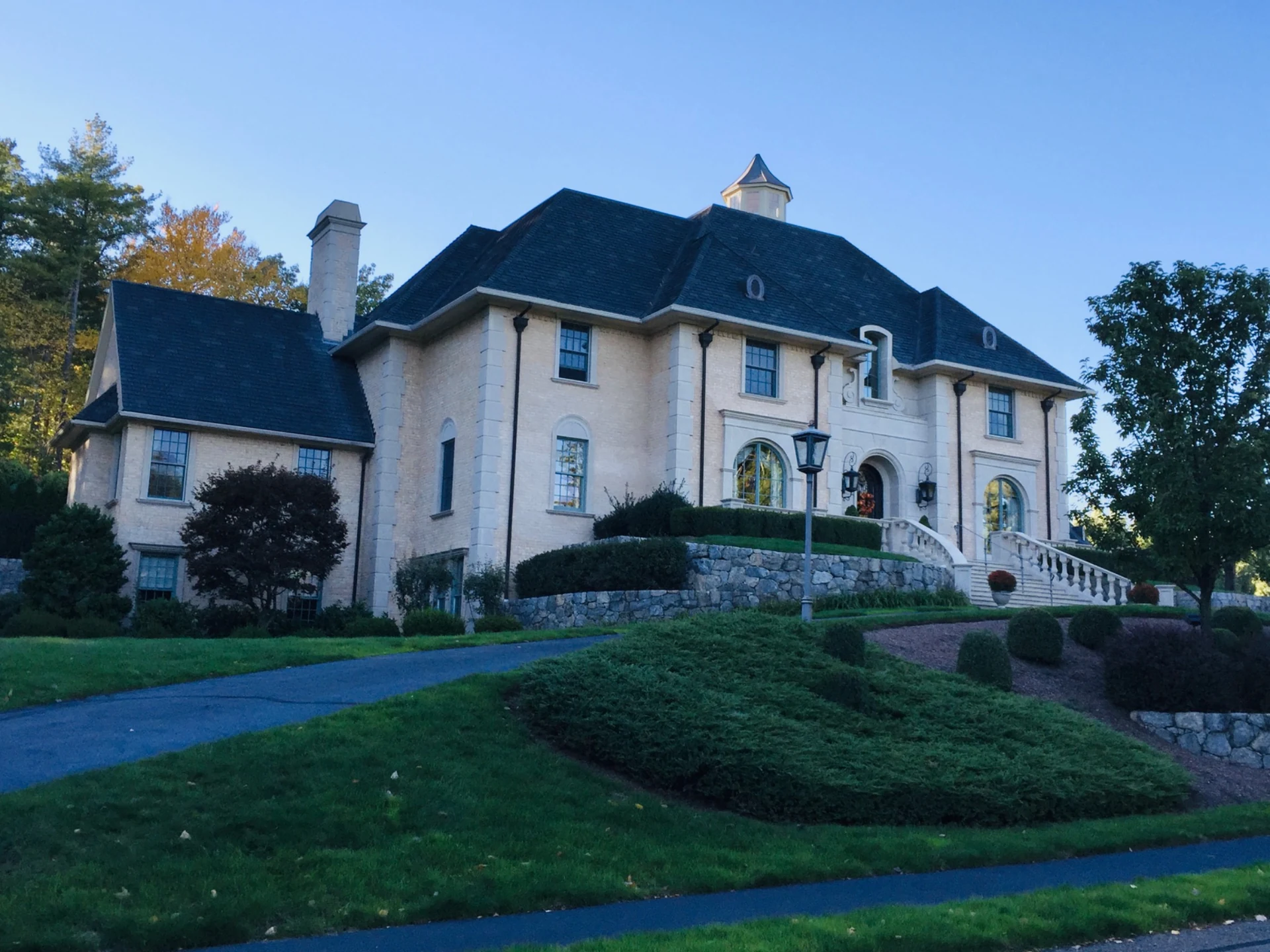 An expansive pale stone home with tall windows, a dark hipped roof, and terraced landscaping